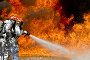 Two firefighters in PPE using a water hose to extinguish a fire, highlighting safe handling and storage of flammable liquids in mines